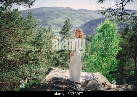 bella giovane sposa dai capelli rossi nella foresta con una corona floreale sulla testa. donna in lungo vestito bianco all'aperto il giorno d'estate. giorno di nozze. Foto Stock