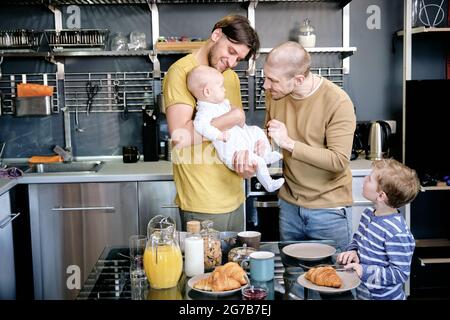 Ragazzo curioso che guarda i suoi padri allegri giocando con il bambino ragazzo in cucina Foto Stock