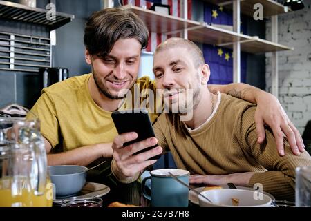 Hugging giovane felice coppia LGBT controllare i social media sullo smartphone insieme, leggere notizie o fare acquisti online Foto Stock