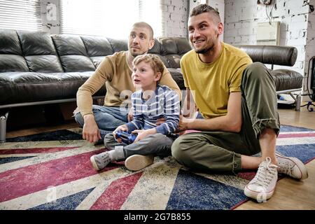 Due padri felici che sostengono il loro figlio piccolo giocando a videogame su console Foto Stock