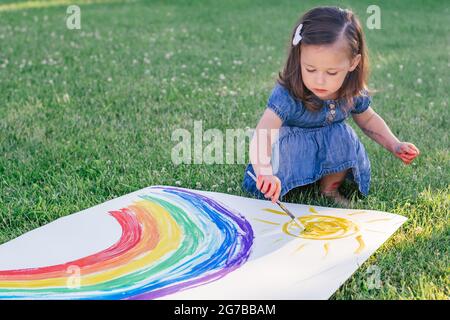 Bambina di 2-4 anni dipinge l'arcobaleno e il sole su un grande foglio di carta seduto su prato verde Foto Stock