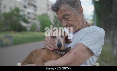 Donna anziana di 90 anni con capelli grigi e profonde rughe si siede all'aperto in una struttura di vita assistita su panca con piccolo cane dachshund. Vecchi abbracci femminili Foto Stock