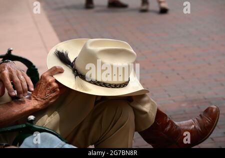 Un uomo che indossa stivali da cowboy e un cappello da cowboy Stetson di paglia si rilassa su una panchina del parco a Santa Fe, New Mexico. Foto Stock