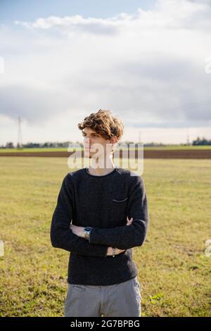 Ritratto di un giovane bel caucasico con le braccia ripiegate in un campo d'erba all'aperto. Ragazzo sicuro e premuroso guarda avanti Foto Stock