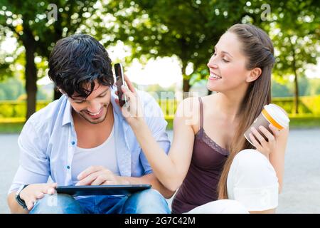 L uomo e la donna o una giovane coppia di bere il caffè e seduta nel parco della città in estate e utilizzo di smart phone e computer tablet Foto Stock