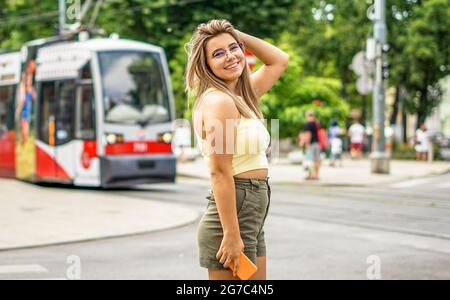 Giovane ragazza sorridente che posa e mette a fuoco la macchina fotografica - Teenager che cammina nella grande città in estate vestito che tiene il suo smartphone Foto Stock