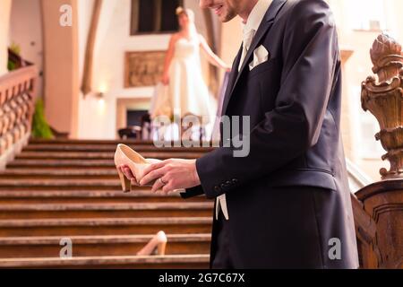Sposo sorridente tenendo alta tallone nella sua mano in piedi di fronte a scale Foto Stock