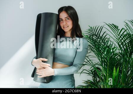 Giovane donna modesta con sorriso sottile che posa con un tappetino yoga, abbracciandolo. Foto Stock