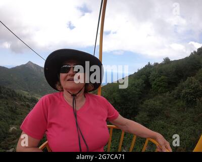 Donna anziana che ama fare un giro in funivia in cestino giallo dalla cima dell'isola d'Elba Foto Stock