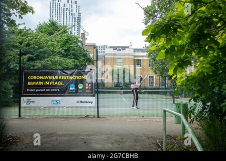 Londra- Luglio, 2021: Persone che giocano a Tennis a Hackney, Londra Est con un grande cartello informativo sul Coronavirus che informa che le restrizioni rimangono in vigore Foto Stock