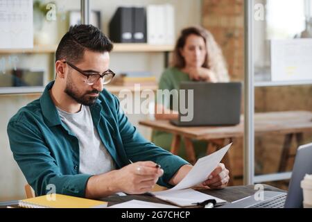 Giovane uomo d'affari serio in occhiali seduto sul suo posto di lavoro di fronte al computer portatile e la lettura di un contratto in ufficio Foto Stock