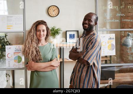 Ritratto di una giovane coppia che sorride alla fotocamera mentre si trova vicino alla parete di vetro con le carte in ufficio Foto Stock