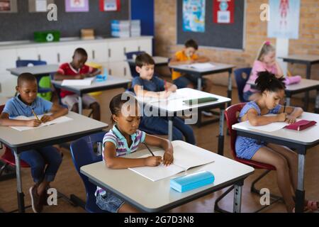 Gruppo di studenti diversi che studiano mentre si siedono sulle loro scrivanie nella classe a scuola Foto Stock