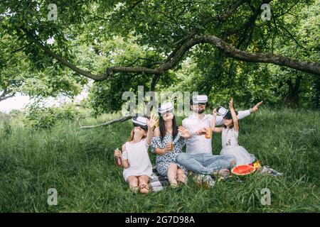 Giovane famiglia caucasica divertirsi in occhiali virtuali all'aria aperta. Felici genitori con due figlie sedute su una morbida coperta e giocando a giochi. Tempo libero e concetto di intrattenimento. Foto Stock