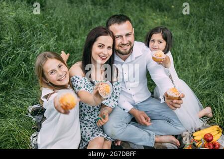 Ritratto di bella famiglia che si posa insieme all'aria fresca con gustose cupcake fatte in casa in mano. Padre, madre e due figlie che hanno pic-nic estivo in giardino verde. Foto Stock