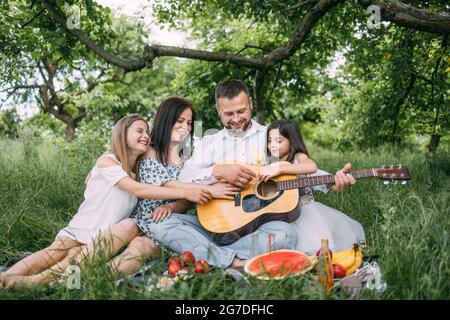 Felice famiglia con le figlie carine che hanno pic-nic nel giardino verde con canzoni di chitarra. I genitori e i due bambini trascorrono il tempo libero insieme al divertimento. Foto Stock