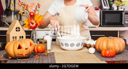 Una donna versa la farina da un bicchiere in pasta per biscotti per Halloween in cucina con un arredamento autunnale. Casa accogliente. Fare biscotti per Halloween. Foto Stock