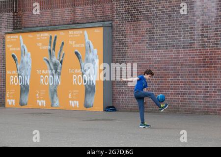 Londra, Regno Unito, 13 luglio 2021: Un ragazzo calcia una palla contro un muro a Tate Modern, si legano a poster per una mostra di sculture di Rodin. I posti per giocare all'aperto sono stati ridotti nel corso degli anni e non tutti i bambini hanno accesso allo spazio giochi. Anna Watson/Alamy. Foto Stock