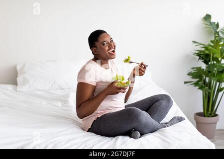 Concetto di dieta snellente bilanciata. Donna nera in sovrappeso che mangia insalata di verdure sul letto a casa, a lunghezza intera Foto Stock