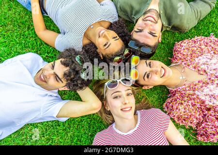 Girato dall'alto di cinque amici multirazziali che si trovano testa a testa con occhiali da sole colorati che guardano la macchina fotografica dal basso. Gara mista di smilin Foto Stock