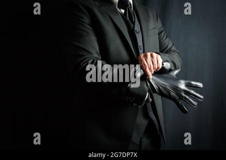 Ritratto di Gentleman in abito scuro che tira sui guanti in pelle nera su sfondo nero. Uomo d'affari elegante vestirsi. Foto Stock