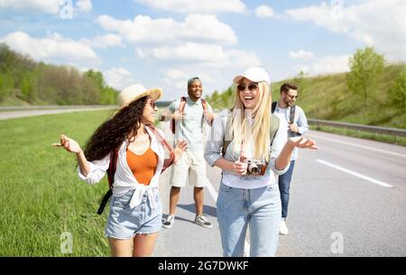 Giovani amici diversi con zaini che camminano lungo la strada, che viaggiano insieme, che hanno un viaggio autosstop Foto Stock