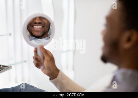 Nuovo sorriso. Uomo nero guardando lo specchio mentre si siede nella sedia dentista Foto Stock