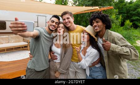 Felici amici diversi che posano vicino camper in campagna, facendo gesti diversi, prendendo selfie sullo smartphone Foto Stock