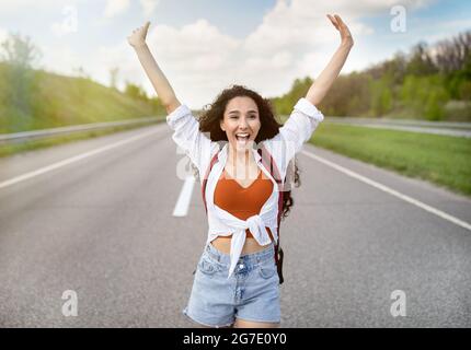 Giovane donna eccitata che cammina lungo l'autostrada, sollevando le mani e gridando, sentendosi felice e libero durante il viaggio estivo Foto Stock