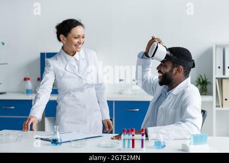 Scienziato afro-americano in piedi vicino a un collega in visore vr vicino alle attrezzature in laboratorio Foto Stock