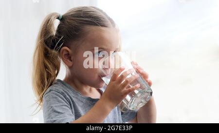 Bambina bionda in abito bianco beve acqua da un bicchiere al chiuso. Il bambino carino sta bevendo una tazza di acqua Foto Stock