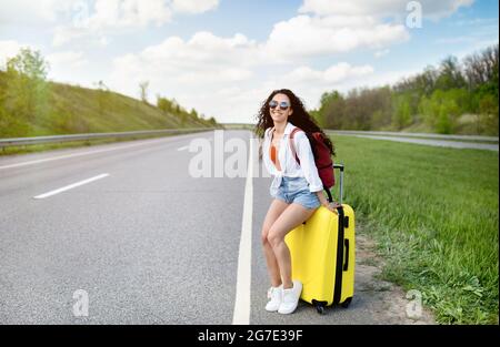 Bella giovane donna seduta sulla valigia lungo la strada, sventolando in auto, hitchtrekking sulla strada, avendo viaggio in estate Foto Stock