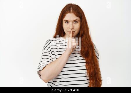 Hush, non dire a nessuno. Misteriosa ragazza rossa che gridava, fare segno shh con il dito sulle labbra e sorridendo, dicendo segreto, chiedendo di tenere tranquillo, in piedi Foto Stock