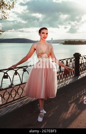 una giovane ed elegante ballerina femminile in un abito rosa con gonna organza piena, una ballerina si pone vicino ad una recinzione in ferro battuto presso la baia del mare ai raggi di Foto Stock