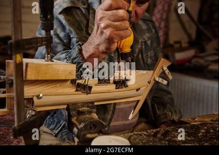 Carpentiere in azione, stili di vita, concetti hobby. Artigiano che attacca dettagli in legno su una barca a vela nella sua propria officina. Foto Stock