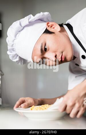 Chef asiatico la placcatura di una ciotola di cibo con attenzione tergi intorno al lato per rimuovere eventuali fuoriuscite, vicino sul suo volto con sguardo di concentrazione Foto Stock