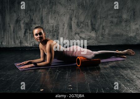 Giovane donna atletica muscolare con un corpo perfetto bello nello sportswear massaggiando i muscoli dei piedi con massaggiatore a rullo in schiuma sdraiato sul tappetino yoga. Foto Stock