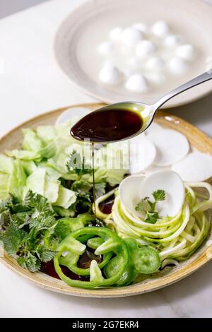 Versando olio di semi di zucca su verdure fresche grezze verdi ed erbe, zucchine di spaghetti, rafano bianco, paprika verde, insalata di ghiaccio, palline di mozzarella per coo Foto Stock