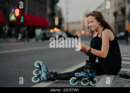 La donna sportiva e felice indossa le lame a rullo sportslothes si siede sui controlli stradali newsfeed via smartphone si rompe dopo che il pattinaggio inline si pone contro Foto Stock