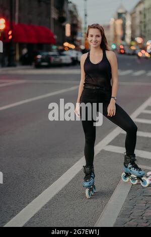 Foto a lunghezza intera di giovane donna attiva in buona forma fisica vestita di activewear nero gode rollerblading durante il buon giorno d'estate pone sopra Foto Stock