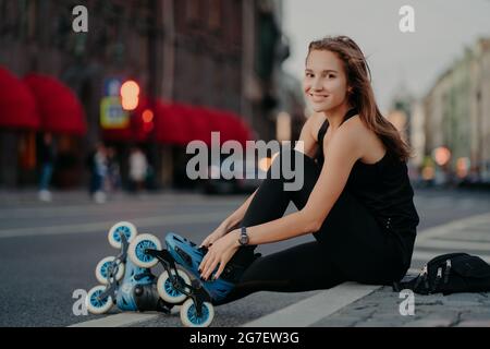 Fit positivo donna mette su rollerblade si siede su asfalto contro sfondo strada offuscato ha espressione positiva va in per lo sport ha all'aperto Foto Stock