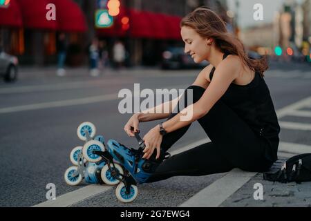 Un tiro all'aperto di lacci attivi si prepara per il giro si siede sulla strada contro lo sfondo della città occupato vestito in nero activewear gode Foto Stock