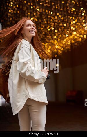 Giovane Hipster femmina in Beige Jacket Walking in City Street. Avere buon umore sopra il Bokeh festoso incandescente. Garlands luci sfondo urbano. Verticale o Foto Stock