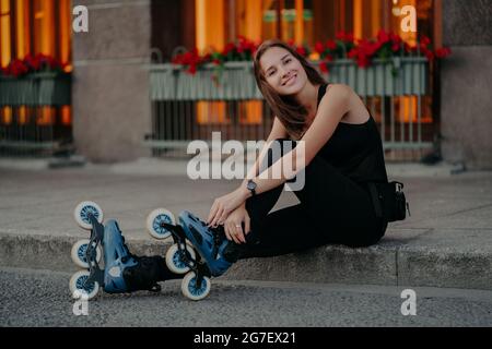 Piacevole aspetto giovane donna europea indossa pattini a rotelle si rompe dopo aver cavalcato pose all'aperto vestita in nero activewear ha sorriso felice sul viso Foto Stock