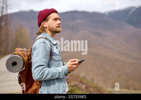 brutale uomo caucasico che viaggia nella natura selvaggia con smartphone, con zaino marrone in pelle, stile autunnale, viaggiatore, avventura, guglie gratis Foto Stock