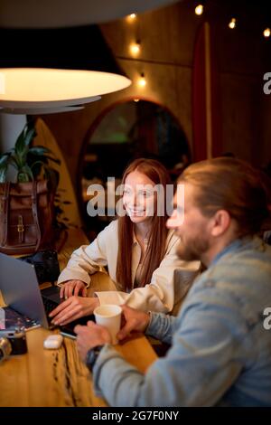 Vista laterale ritratto di allegri manager caucasici fotografi rosso testa femmina e ragazzo bearded che lavorano in caffetteria, utilizzare il laptop, navigare in internet, avendo t Foto Stock