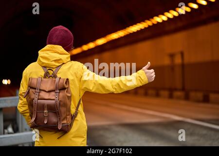 Vista dal retro su hitchhiking giovane uomo intorno al paese, cercando di prendere l'auto di passaggio per viaggiare. Uomo caucasico hipster in camice giallo con dorso Foto Stock