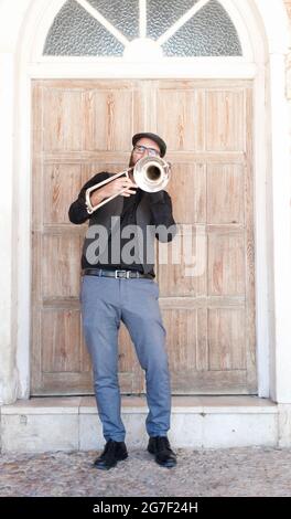 Musicista jazz con occhiali e barba che suona il trombone in piedi per strada. Vestito con un cappello inglese, una giacca, pantaloni blu. Dietro è un d di legno Foto Stock