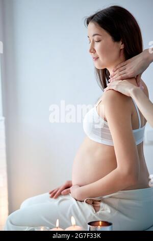 Bella donna incinta asiatica ottenere massaggio spa sulla schiena, si siede tinta, in bianco reggiseno. Vista laterale ritratto di bella signora in salone spa in luce Foto Stock