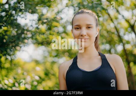 Bella donna sportiva in abbigliamento sportivo utilizza auricolari wireless per ascoltare la musica riposa nel parco verde guarda direttamente la fotocamera ha riposo dopo l'esterno Foto Stock